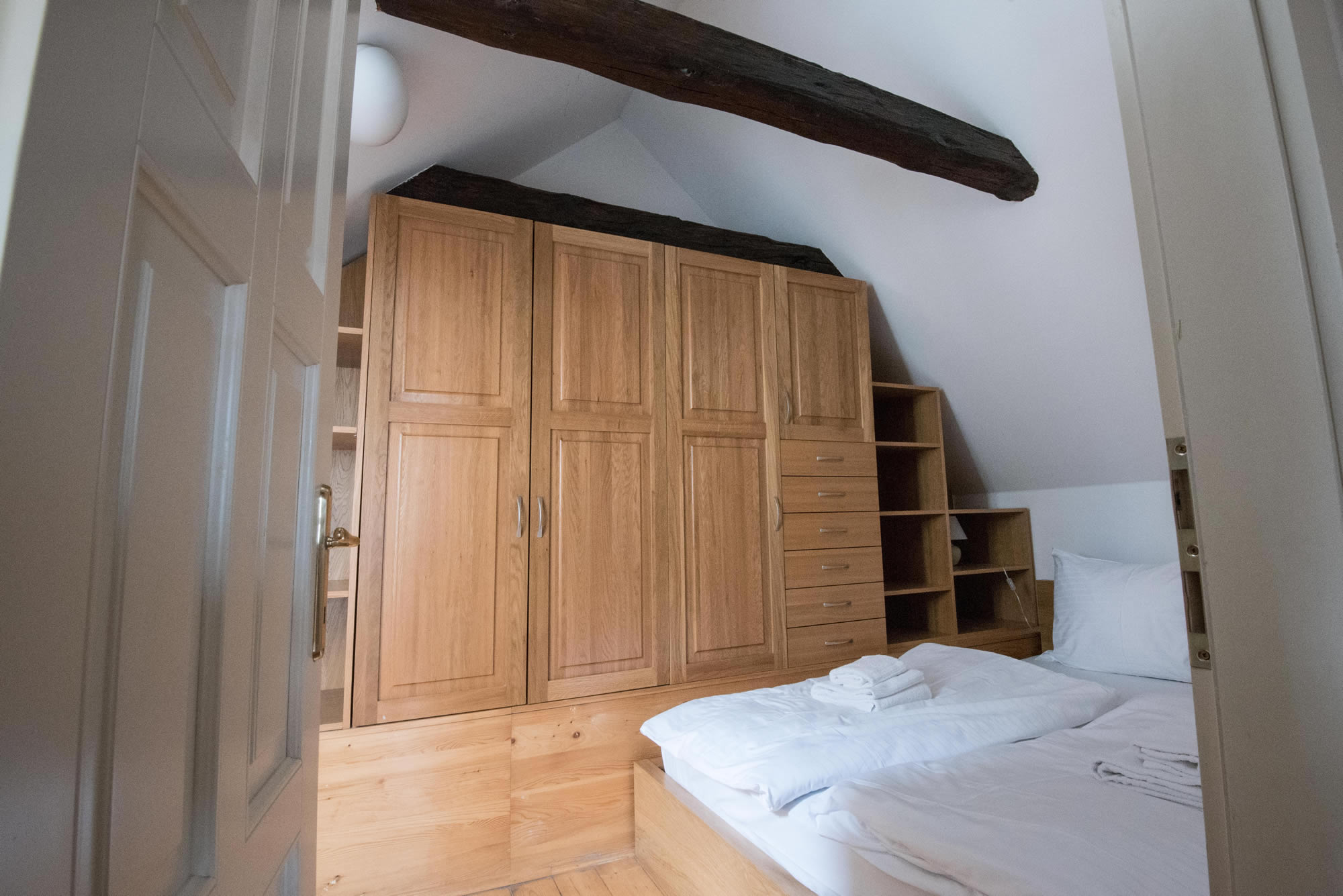 Attic bedroom with white bed sheets, wooden wardrobe and wood beams.