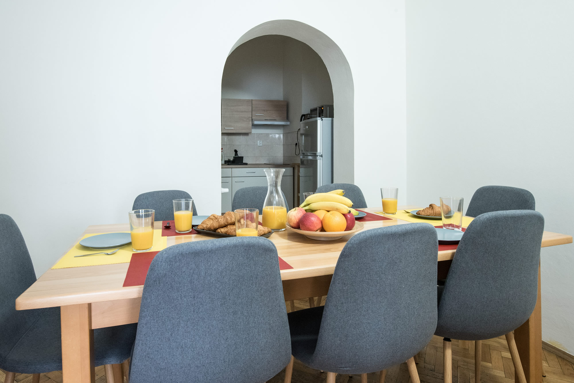 Dining table with fruit, juice and croissant. Grey chairs are around. There is a kitchen behind.