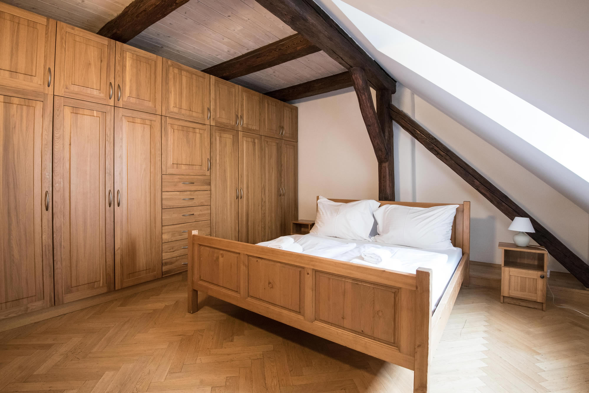 Bedroom with double bed. Wooden beam are in the attic room. Parquet on the floor and large wooden closet on the wall.