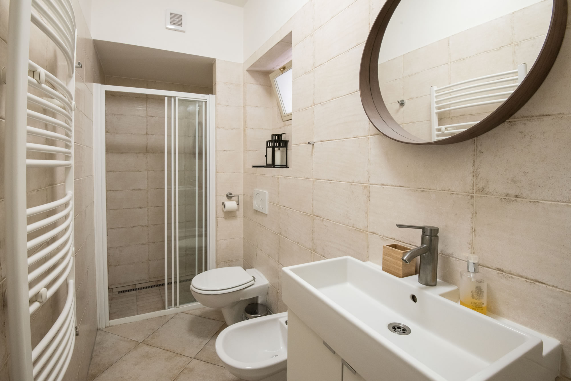 Bathroom with beige tiles on the floor and on the walls. A shower, bidet, toilet and white sink with mirror are in the room.