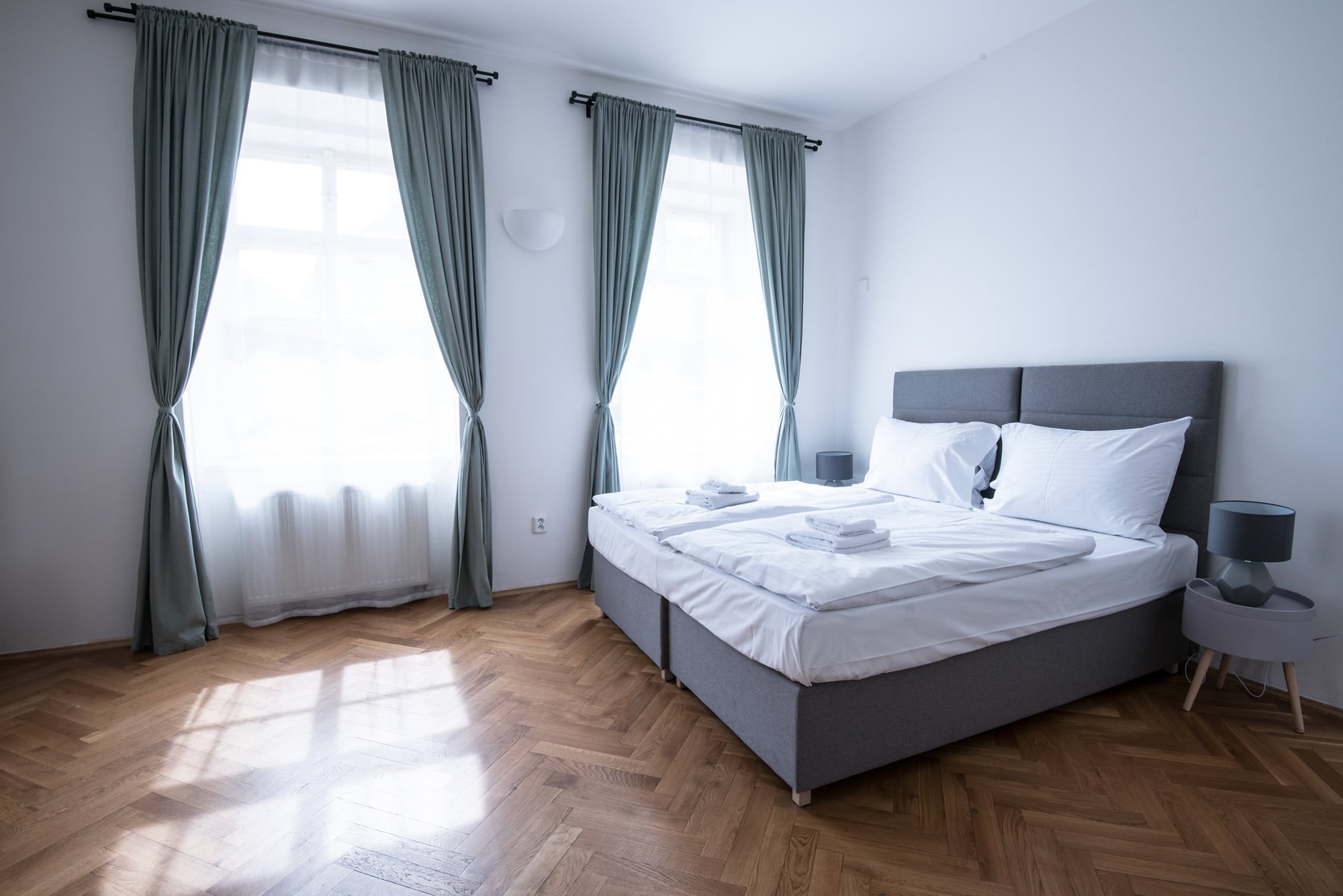 Bedroom with grey king sized bed and white bed sheets. Wooden parquet is on the floor, grey curtains are by the windows.