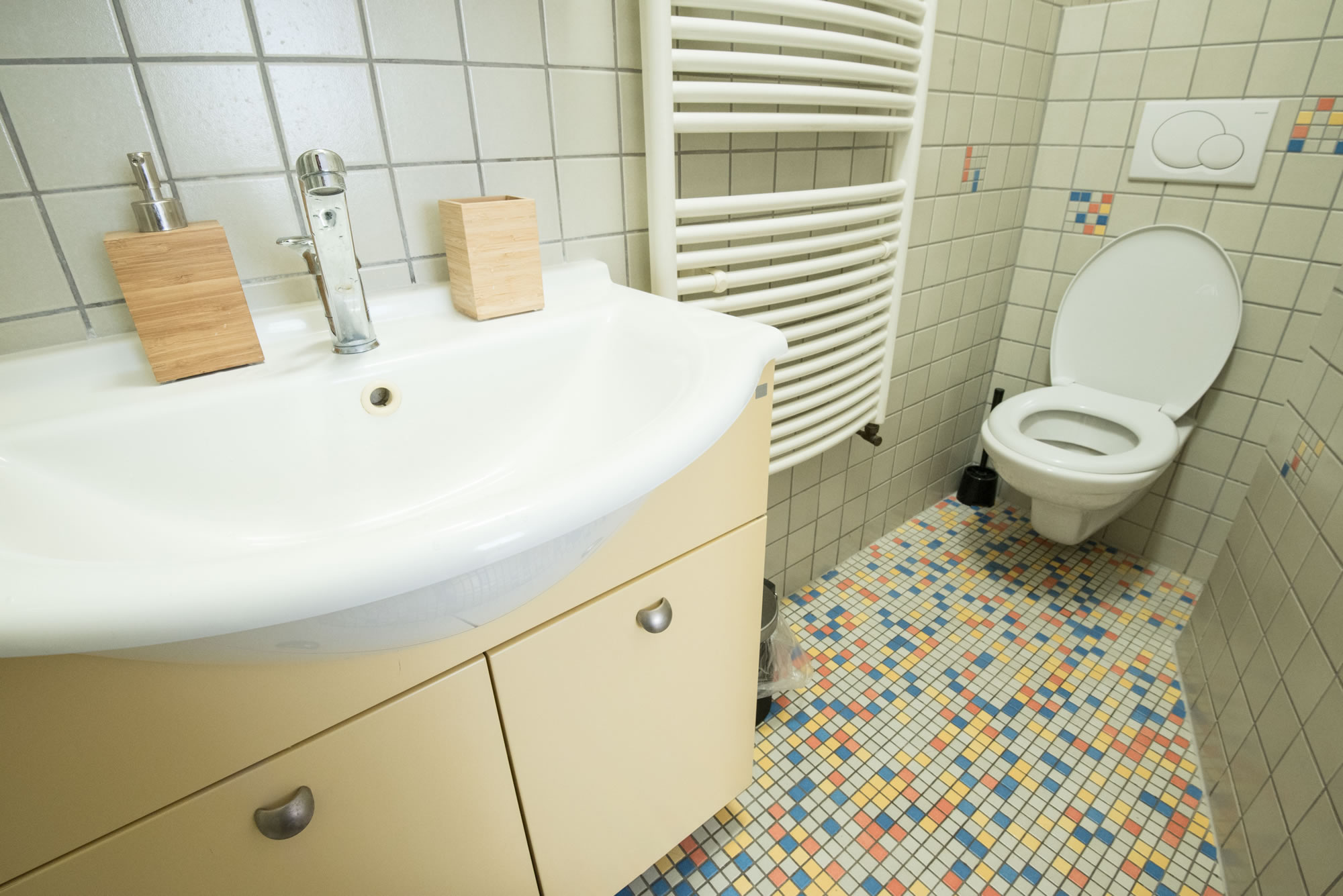 Bathroom with beige tiles on the wall and colored tiles on the floor. There is a toilet and a sink in the bathroom.
